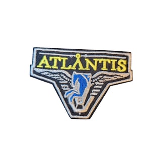 May include: Embroidered patch with the word "ATLANTIS" in yellow letters. The patch features a blue Pegasus design with wings, set against a black background with white and yellow accents. The patch has a rectangular shape.