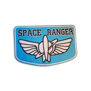 Op de afbeelding: Een rechthoekige geborduurde patch met afgeronde hoeken, een lichtblauwe achtergrond en een witte rand. De tekst SPACE RANGER is in zwarte hoofdletters geborduurd boven een wit gevleugeld embleem met een centrale verticale raketvorm.