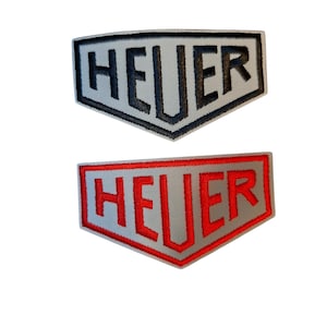 Puede incluir: Dos parches bordados en forma de escudo con la palabra HEUER en letras mayúsculas. El parche superior tiene un fondo gris claro con letras y borde negros. El parche inferior tiene un fondo gris claro con letras y borde rojos.
