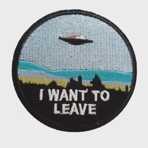 Könnte beinhalten: Runder, gestickter Aufnäher mit schwarzem Rand. Er zeigt eine Landschaft mit einem UFO am Himmel. Der Himmel ist hellblau, darunter eine türkis-gelbe Landschaft. Der weiße Text lautet "I WANT TO LEAVE".