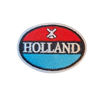 May include: An embroidered patch with an oval shape. The top half is red, the bottom half is light blue, and the word "HOLLAND" is in white letters. A white cross-shaped design is at the top.