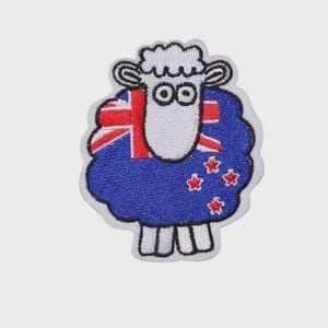 Könnte beinhalten: Gestickter Aufnäher mit einem Schaf mit weißem Gesicht und Beinen, blauer Wolle und der Flagge Neuseelands. Der Aufnäher hat einen weißen Rand und ist mit cartoonartigen Merkmalen gestaltet.