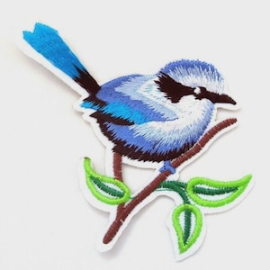 May include: Embroidered patch featuring a blue bird perched on a brown branch with green leaves. The bird has a blue body, a black beak, and a white belly. The patch has a white border.