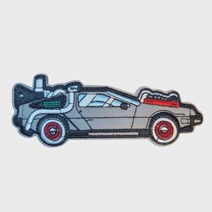 Puede incluir: Parche bordado que representa un coche DeLorean gris con ruedas rojas y detalles del motor rojos. El parche tiene un borde negro y está diseñado para planchar o coser sobre tela. El coche tiene un diseño único con puertas de ala de gaviota.