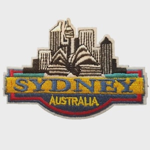 May include: Embroidered patch depicting the Sydney skyline, including the Opera House and skyscrapers. The patch has a yellow border with the word "SYDNEY" in blue and "AUSTRALIA" below. The patch is rectangular.