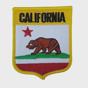 Könnte beinhalten: Gestickter Aufnäher mit dem Design der kalifornischen Staatsflagge. Der Aufnäher hat einen gelben Rand und einen oberen Bereich mit dem Wort "CALIFORNIA". Darunter befindet sich ein weißes Feld mit einem roten Stern, einem braunen Bären, grünem Gras und einem roten Boden.