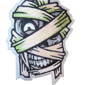 Peut inclure: Écusson brodé représentant une tête de momie de dessin animé avec des bandages. Le motif comprend un œil détaillé, un large sourire et des bandages verts et blanc cassé. L'écusson a une bordure blanche.