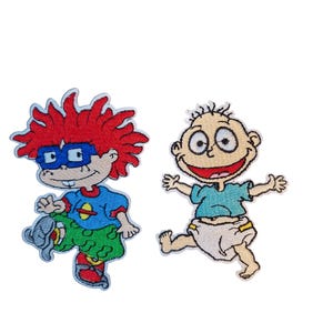 May include: Two embroidered patches featuring cartoon characters. One patch depicts a character with red hair, blue glasses, a blue and green outfit, and a gray shoe. The other patch shows a smiling baby in a blue shirt and diaper.
