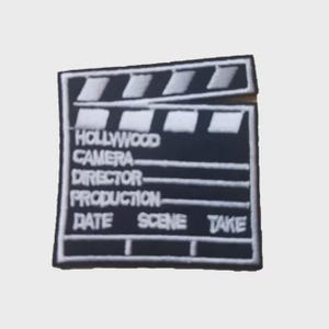 Puede incluir: Un parche bordado en blanco y negro con forma de claqueta de cine. El texto en la claqueta dice "HOLLYWOOD CAMERA DIRECTOR PRODUCTION DATE SCENE TAKE". La claqueta tiene rayas blancas en la parte superior.