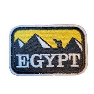 Puede incluir: Parche bordado con un cielo amarillo y pirámides negras con silueta de camello. La palabra "EGYPT" está bordada en letras blancas sobre fondo negro, contorneada en blanco. El parche tiene un borde blanco.