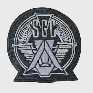 May include: A circular embroidered patch featuring a black background with a silver-toned design. The emblem displays the letters SGC at the top, centred above a stylised triangular shape with horizontal wing-like extensions and circular borders.
