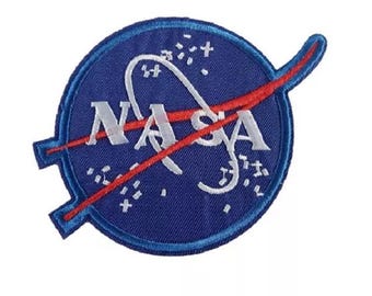 Può includere: Una toppa blu circolare con il logo NASA. Il logo include la parola "NASA" in lettere maiuscole bianche, un percorso orbitale bianco e un trattino rosso. Piccole stelle bianche sono sparse intorno al logo.