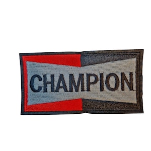 May include: Embroidered rectangular patch featuring the word "CHAMPION" in black on a grey background. The patch has a red and black border.