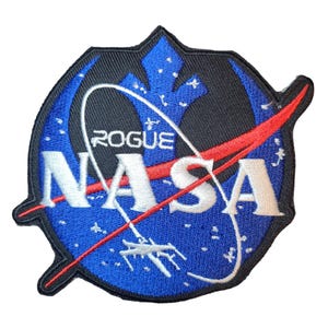 Pode incluir: Patch bordado com um design azul e preto, com a palavra "NASA" em branco. O patch inclui a palavra "ROGUE" e uma linha vermelha no centro. O patch tem uma forma única com bordas pontiagudas.