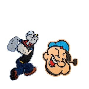 May include: Two embroidered fabric patches featuring the cartoon character Popeye. One patch shows the full figure of the sailor in a black shirt and blue trousers, while the other patch depicts a close-up of the character's face with a pipe.