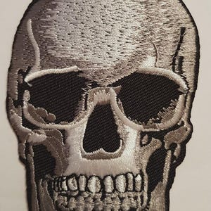 May include: Embroidered skull patch in shades of gray and black. The skull features detailed stitching, with black eye sockets and a detailed jawline. The patch is likely for jackets, bags, or other apparel.
