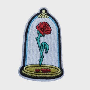 Può includere: Una toppa ricamata raffigurante una rosa rossa con stelo e foglie verdi, racchiusa all'interno di una cupola di vetro azzurro. La cupola poggia su una base dorata con due piccoli petali rossi che riposano sulla superficie all'interno.
