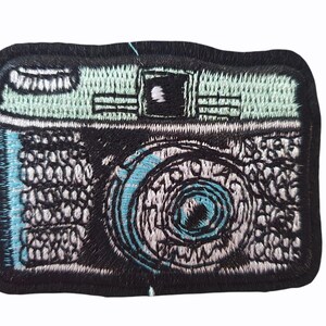 Peut inclure: Écusson brodé représentant un appareil photo vintage. Le patch a une bordure noire, avec le corps de l'appareil photo en noir et blanc, et des accents bleu clair et sarcelle. L'objectif présente un motif en spirale.