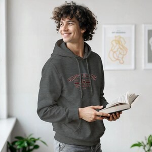 Peut inclure: Un sweat à capuche gris anthracite avec le texte "KAST KULTUR EST. 16 OVERALL" en bordeaux. Le sweat à capuche a une capuche à cordon et une poche avant. La personne tient un livre.