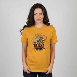 May include: Mustard yellow t-shirt featuring a detailed graphic of a tree with exposed roots and branches. The text "FUCKING" is printed across the trunk in a bold, stylized font. A red sun-like shape is behind the tree.