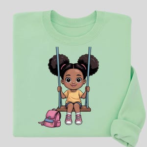 Può includere: Felpa verde chiaro con un'illustrazione a cartoni animati di una bambina su un'altalena. La bambina ha i capelli scuri in due chignon, indossa una maglietta gialla, pantaloncini blu e scarpe rosa. Uno zaino rosa è a terra.