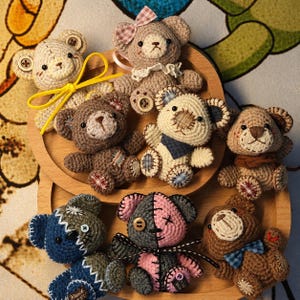May include: A selection of handmade crochet teddy bears in shades of brown, beige, blue, and pink. Each bear features unique details such as button eyes, stitched patches, and bows. The bears are displayed on a wooden tray, highlighting their handcrafted quality.