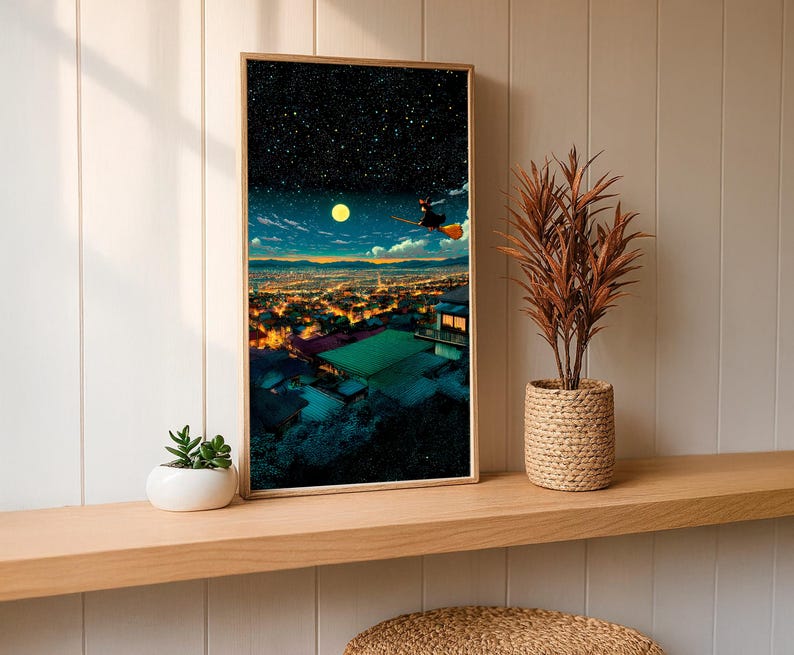 May include: A framed art print depicts a night scene with a witch flying on a broom over a city. The artwork features a starry sky, a full moon, and city lights. The print is displayed on a wooden shelf with a plant in a woven pot.