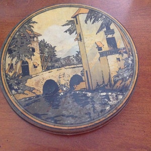 May include: Round, vintage tin with a painted scene of a bridge, buildings, and palm trees. The artwork features muted colors of tan, brown, and gold, with a dark border. The tin is set on a wooden surface.