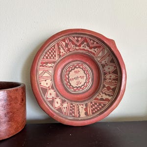 May include: A handmade, red ceramic plate with a detailed geometric pattern in white and brown. The plate has a circular shape with a slightly raised rim and a small handle. The center of the plate features an inscription.