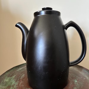 May include: A matte black ceramic teapot with a smooth, rounded body, a curved spout, a sturdy side handle, and a matching lid with a small knob. The teapot features a solid, dark finish and a simple, functional design for serving hot beverages.