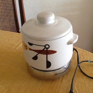 May include: White ceramic slow cooker with a black and brown design of a stylized bird with a spoon in its beak. The cooker has a chrome base and a cord.