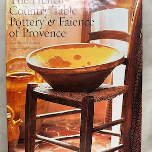 May include: A hardcover book titled The French Country Table Pottery & Faience of Provence, featuring text by Bernard Duplessy and photographs by Camille Moirenc. The cover displays a yellow and brown ceramic bowl resting on a wooden chair.
