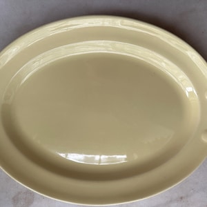May include: A pale yellow oval serving platter with a smooth, glossy finish. The platter features a wide, flat central area and a raised rim with two integrated handles on each side, each accented by two small, circular decorative indentations.