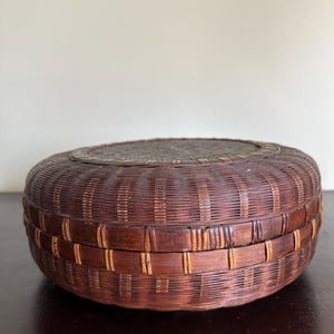 May include: A round, woven basket with a lid. The basket is made of dark brown and tan-colored woven material. The lid is a darker shade of brown and has a woven pattern. The basket is sitting on a dark surface.