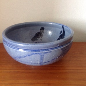 May include: A handmade, blue ceramic bowl with a rounded shape. The bowl features a textured surface and is decorated with dark blue abstract designs. The rim has two dark blue stripes. The bowl is sitting on a wooden surface.