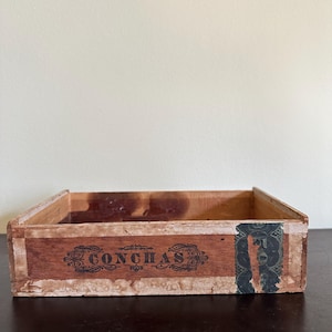 May include: A vintage wooden box with the word "CONCHAS" printed in black script. The box is rectangular with a worn, aged appearance, showing signs of use and age. The box has a dark brown color with a light-colored trim.