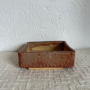 May include: A rectangular ceramic dish with a textured, diamond-patterned exterior in shades of brown and red. The interior has a light yellow hue. The dish sits on a beige base, suitable for serving or display.
