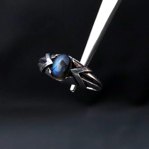 Könnte beinhalten: Ein silberner Ring mit einem blauen Edelstein, gehalten von einer Pinzette vor schwarzem Hintergrund. Der Ring hat ein geometrisches Design mit einem oval geformten Mittelstein. Das Metall des Rings hat eine dunkle, oxidierte Oberfläche.