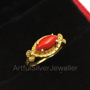 Può includere: Un anello color oro con una gemma rossa brillante di forma ovale. L'anello è incastonato in una fascia decorativa in stile filigrana. È presentato in un portagioie di velluto nero con interno rosso. Il testo "ArtfulSilverJeweller" è visibile.