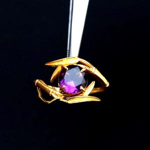 May include: A gold ring with a large, oval, purple gemstone held in place by ornate gold prongs. The ring is held by a pair of tweezers against a black background, highlighting the ring's intricate design and the gemstone's color.