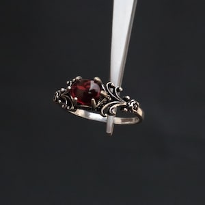 Könnte beinhalten: Ein silberner Ring mit einem dunkelroten Edelstein, gehalten von einer Pinzette vor dunklem Hintergrund. Der Ring hat ein verziertes, florales Design um den Edelstein.
