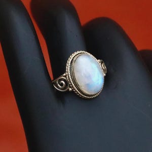 May include: Oval-shaped moonstone ring with a gold-colored band. The moonstone has a pearly white color with iridescent blue and yellow hues. The band features a twisted rope design around the stone and decorative swirls on the sides.