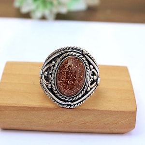 Puede incluir: Un anillo plateado con una piedra preciosa ovalada, moteada y marrón. El anillo tiene un diseño ornamentado con un borde en forma de cuerda y detalles decorativos. El anillo se exhibe sobre un pequeño bloque de madera marrón claro.