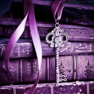 Puede incluir: Una llave decorativa plateada con un diseño ornamentado cuelga de una cinta morada, sobre un fondo de libros antiguos. La llave presenta detalles intrincados y una estética vintage, evocando una sensación de historia y misterio.