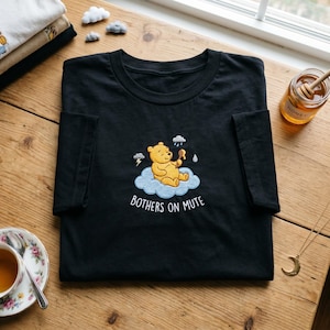 Könnte beinhalten: Schwarzes T-Shirt mit einem gestickten Design eines gelben Bären auf einer blauen Wolke, der Honig hält. Der Text "BOTHERS ON MUTE" befindet sich unter dem Bären. Ein Honigglas und eine Tasse Tee sind ebenfalls zu sehen.
