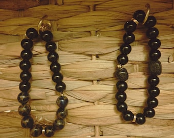 Puede incluir: Dos pulseras de cuentas negras y redondas. Una pulsera tiene algunas cuentas moteadas y detalles dorados. La otra pulsera tiene una cuenta texturizada y detalles dorados. Las pulseras se exhiben sobre una superficie tejida.