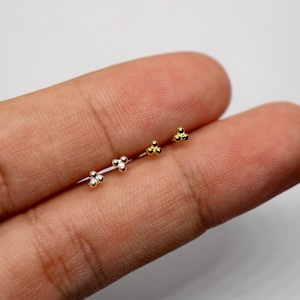 May include: Four pairs of small stud earrings in silver and gold tones. Each earring features a cluster of three tiny spheres. The earrings are displayed on a light background.