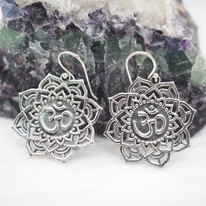May include: A pair of silver earrings with a mandala design. Each earring features an Om symbol at the centre, surrounded by detailed floral patterns. The earrings have a hook closure and are set against a purple and green stone background.