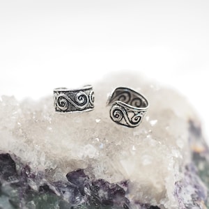 May include: Two silver-toned ear cuffs with an intricate, swirling design. The cuffs are displayed on a light-colored, textured surface. The ear cuffs have a vintage or antique aesthetic.