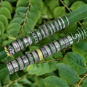Op de afbeelding: Een assortiment zilveren ringen met diverse ontwerpen, waaronder gevlochten, bloemen- en geometrische patronen, gepresenteerd op een groen blad. Er is ook een enkele gouden ring zichtbaar. De ringen zijn geplaatst tegen een achtergrond van groene bladeren.